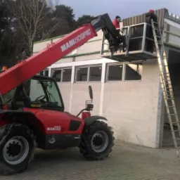 Czerwony podnośnik teleskopowy Maniscopic przy budowie hali. Pracownicy na platformie montują elementy konstrukcyjne. Widoczna drabina oparta o budynek. Hala w stanie surowym.