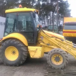 Żółta koparko-ładowarka na placu budowy, z widoczną kabiną i łyżką. W tle ciężarówka MAN i inne maszyny. Teren ziemny, drzewa w tle.