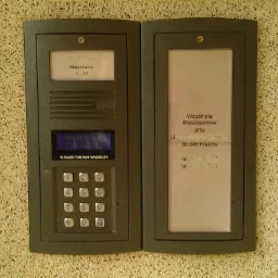 Szary dwuczęściowy panel domofonu z wyświetlaczem LCD i klawiaturą numeryczną zamontowany na teksturowanej ścianie. Wyświetlacz pokazuje komunikat 'WYBIERZ NR I POCZEKAJ'. Druga część zawiera...