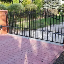 Czarna, metalowa brama dwuskrzydłowa z automatyką, zamontowana na ceglanych słupkach, na posesji z kostką brukową i widocznym samochodem dostawczym w tle.