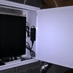 Otwarta biała szafka z zamkiem, zawierająca czarny rejestrator, router AirLive z diodami LED i zasilacz, zamontowana na drewnianej płycie w pomieszczeniu z widocznym fragmentem stropu.