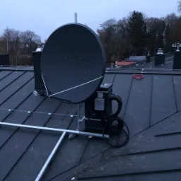 Antena satelitarna zamontowana na metalowej konstrukcji na dachu pokrytym blachą, widoczne przewody i elementy mocujące, w tle drzewa i fragmenty budynków w pochmurny dzień.