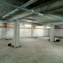 YouLight Julia Wawrzyniak - Wnętrze surowego pomieszczenia z widocznymi instalacjami elektrycznymi pod sufitem, rurami w kolorze pomarańczowym i czerwonym, betonowymi słupami oraz oświetleniem ledowym.