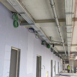 YouLight Julia Wawrzyniak - Wnętrze budynku w trakcie prac instalacyjnych: widoczne koryta kablowe pod sufitem, rury na podłodze i pracownik na drabinie w oddali.