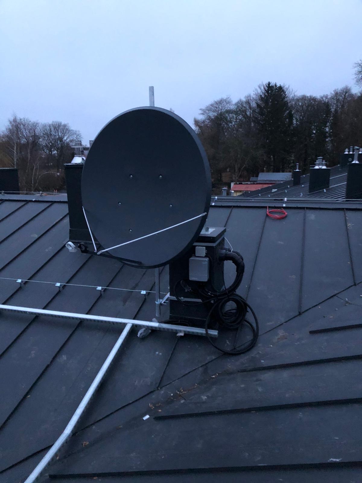 Antena satelitarna zamontowana na metalowej konstrukcji na dachu pokrytym blachą, widoczne przewody i elementy mocujące, w tle drzewa i fragmenty budynków w pochmurny dzień.
