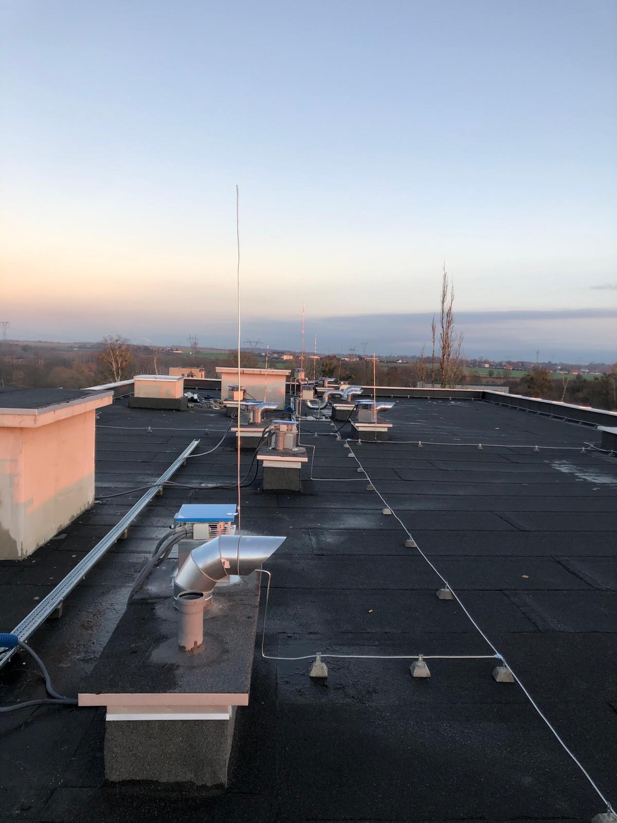 Instalacja odgromowa na płaskim dachu budynku, widoczne metalowe maszty i przewody uziemiające, w tle krajobraz z drzewami i polami o zmierzchu.