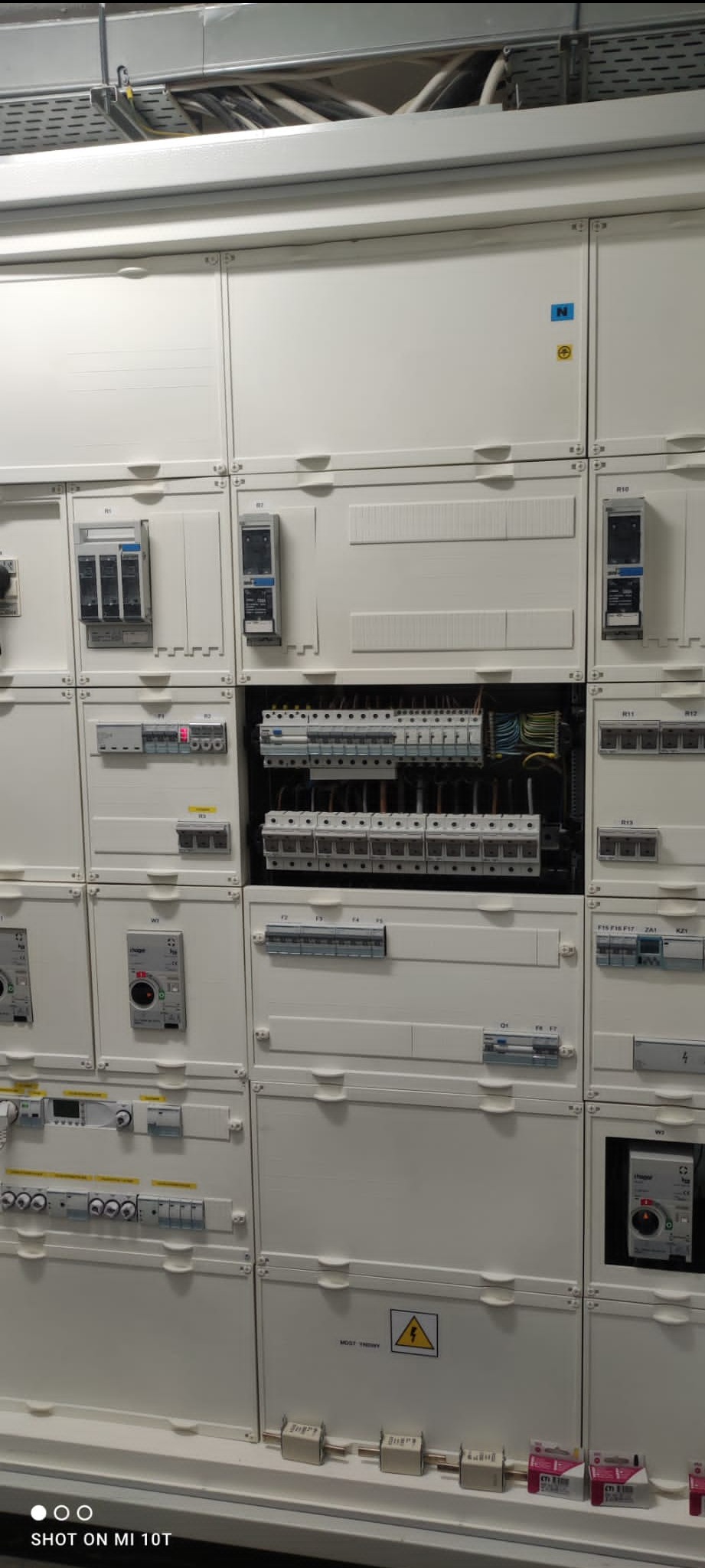 Rozdzielnica elektryczna z widocznymi bezpiecznikami, wyłącznikami i przewodami, umieszczona w białej obudowie z modułowymi panelami.