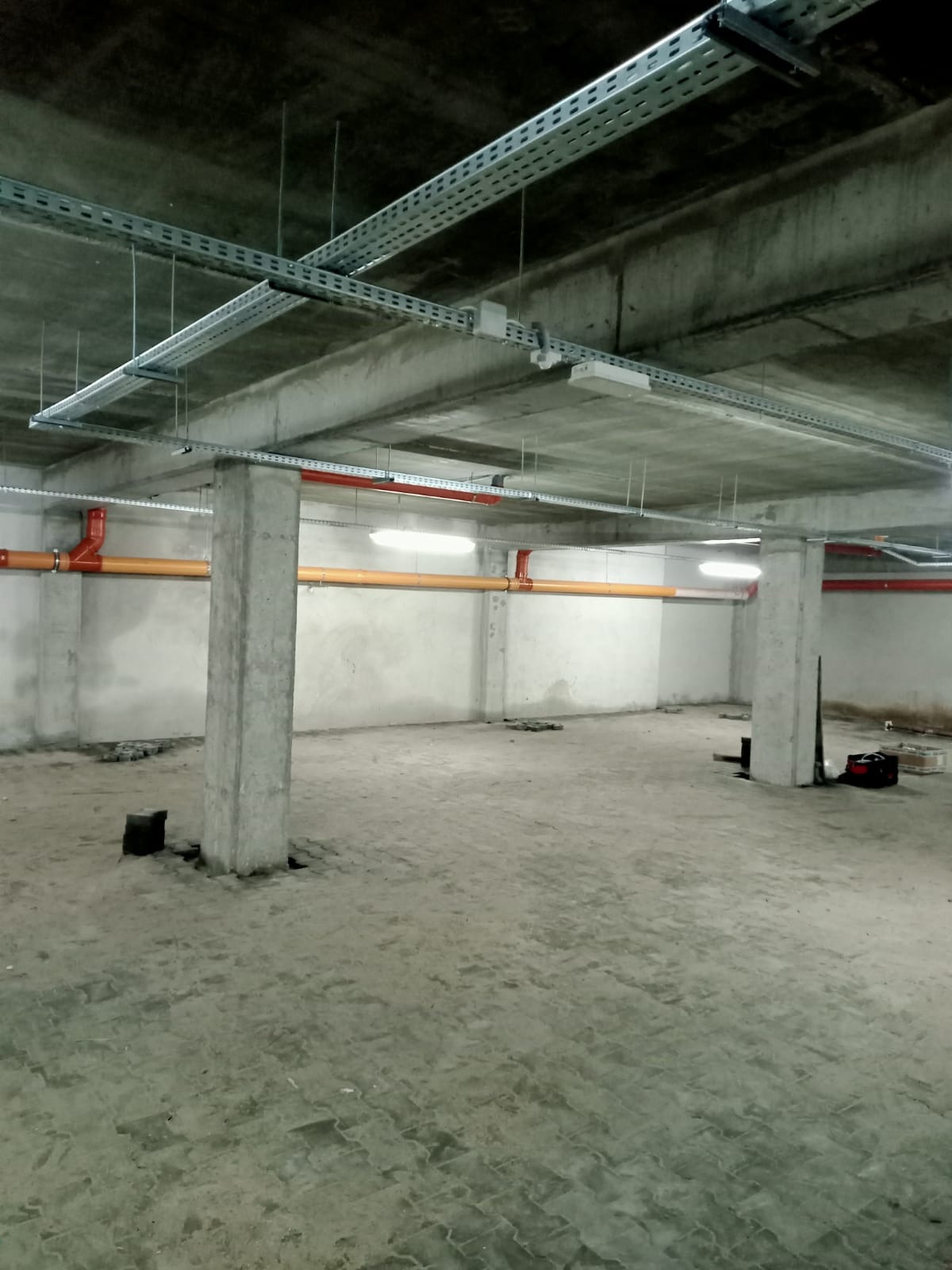 Wnętrze surowego pomieszczenia z widocznymi instalacjami elektrycznymi pod sufitem, rurami w kolorze pomarańczowym i czerwonym, betonowymi słupami oraz oświetleniem ledowym.