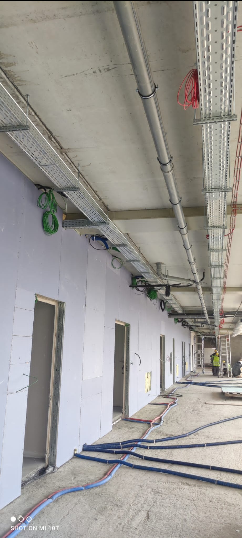 Wnętrze budynku w trakcie prac instalacyjnych: widoczne koryta kablowe pod sufitem, rury na podłodze i pracownik na drabinie w oddali.