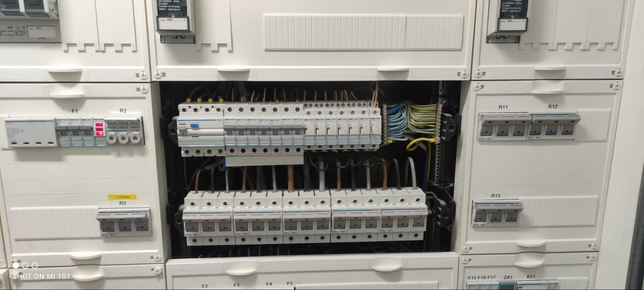 Otwarta skrzynka rozdzielcza z bezpiecznikami, widoczne różnokolorowe przewody i wyłączniki, niektóre z nich w pozycji włączonej, inne wyłączone, nad bezpiecznikami naklejki z oznaczeniami.