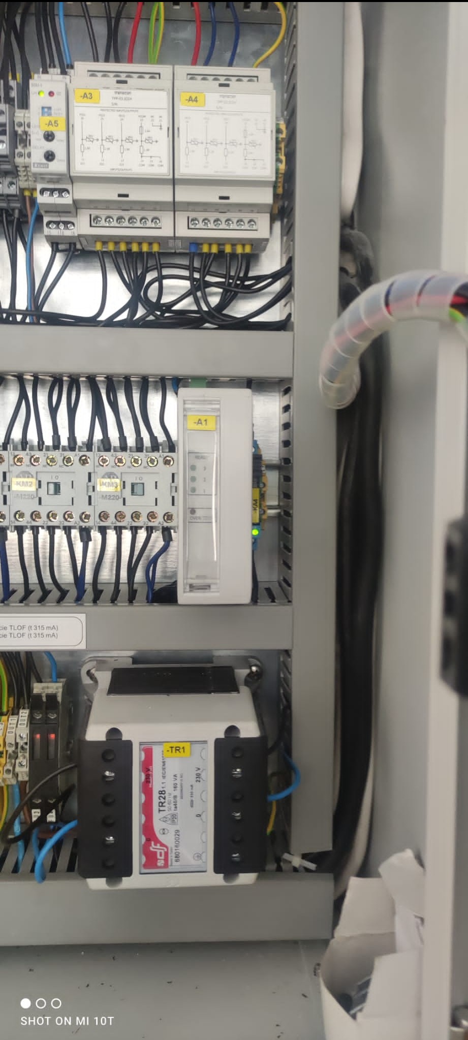 Wnętrze szafy sterowniczej z widocznymi modułami zabezpieczeń, przekaźnikami i okablowaniem. Widoczne oznaczenia A1, A3, A4, A5 oraz TR1 na elementach. Kabel spiralny w prawym górnym rogu.