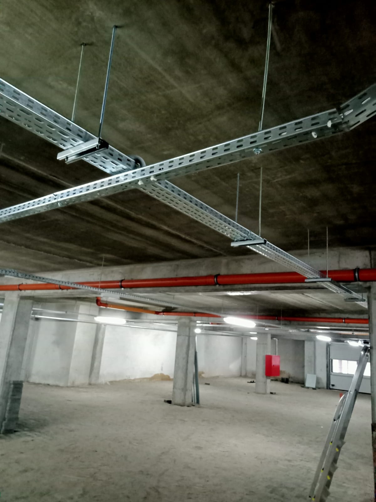 Metalowe korytka kablowe podwieszone do sufitu w surowym stanie, widoczne również pomarańczowe rury instalacyjne, betonowe słupy i drabina oparta o ścianę w tle.