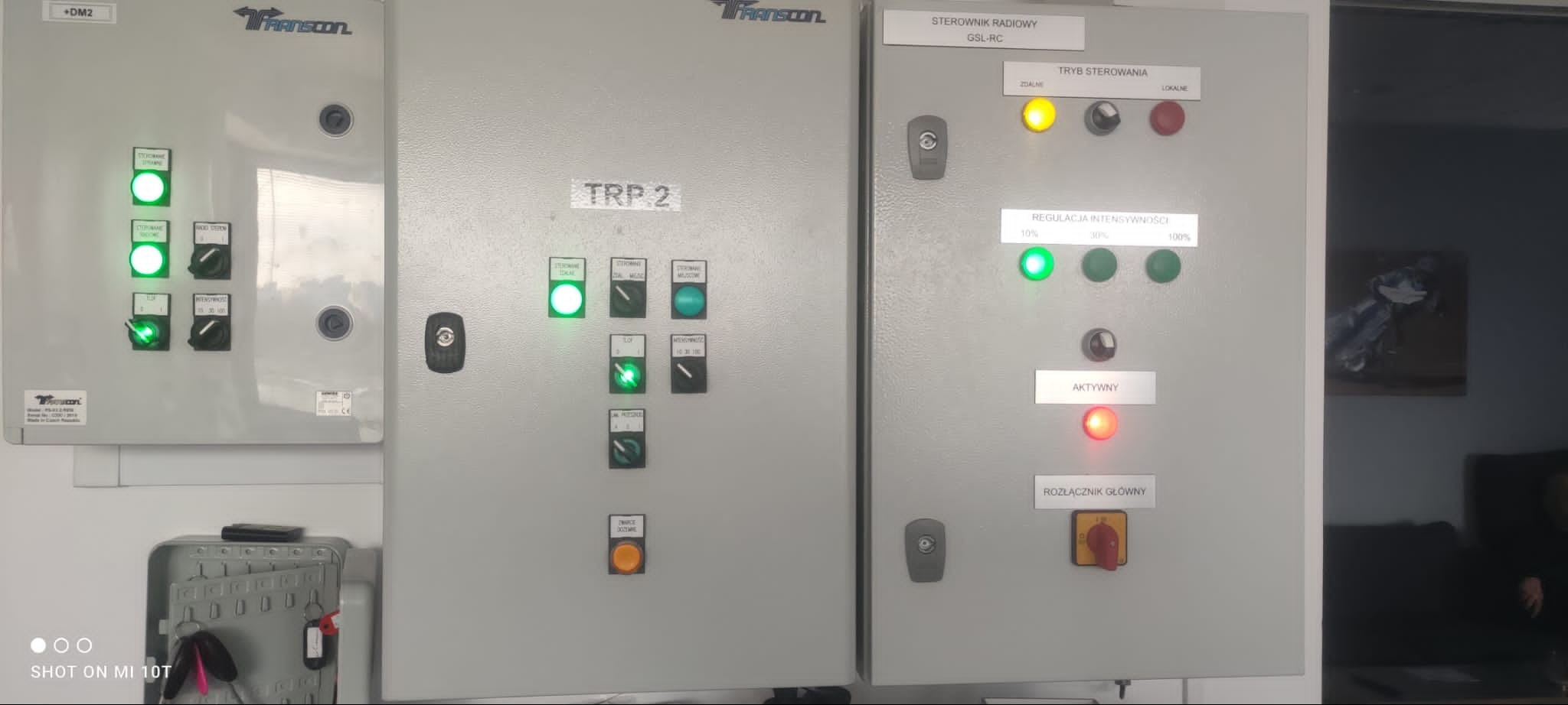 Trzy szare szafy sterownicze z widocznymi panelami kontrolnymi, diodami sygnalizacyjnymi i przełącznikami, z logo Transcon na górze.