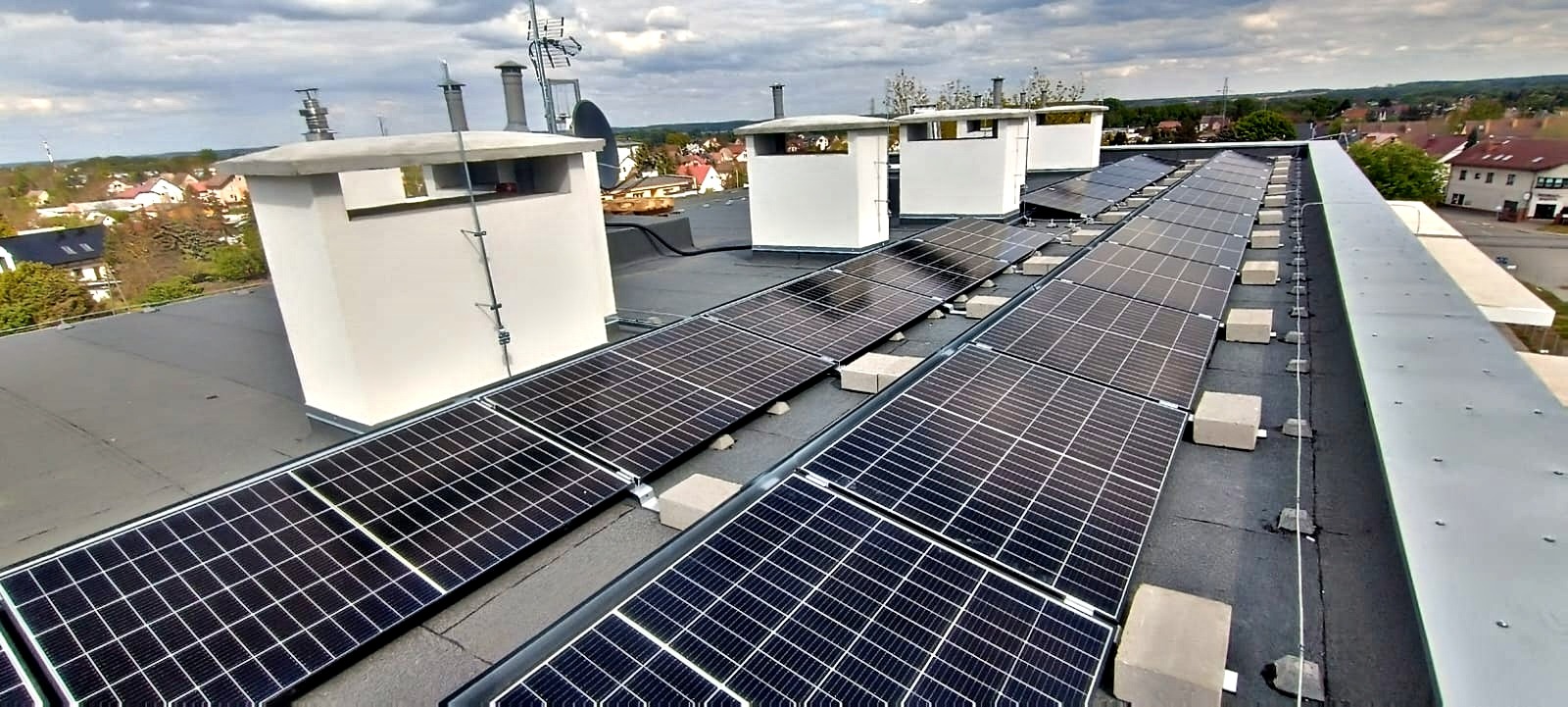 Panele fotowoltaiczne zamontowane na płaskim dachu budynku, widok z góry. Na dachu kominy wentylacyjne. W tle zabudowa miejska i pochmurne niebo.