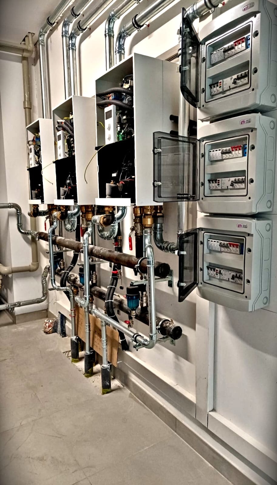 Rozbudowana instalacja z kotłami i rozdzielnicami elektrycznymi w pomieszczeniu technicznym, widoczne rury i kable. Otwarte skrzynki z bezpiecznikami.