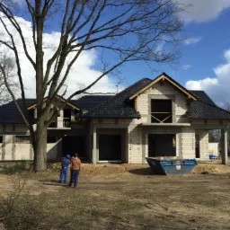 Budowa domu jednorodzinnego z ciemnym dachem, widoczne bloczki i okna bez szyb, na pierwszym planie kontener budowlany i dwie osoby. Widok z zewnątrz, dzień.