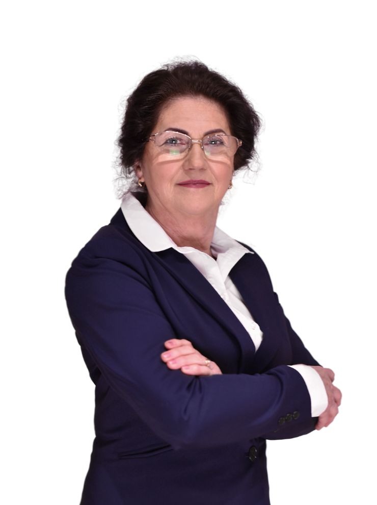 Portret kobiety w okularach, ubrana w granatowy żakiet i białą koszulę, pozująca z założonymi rękami na białym tle.