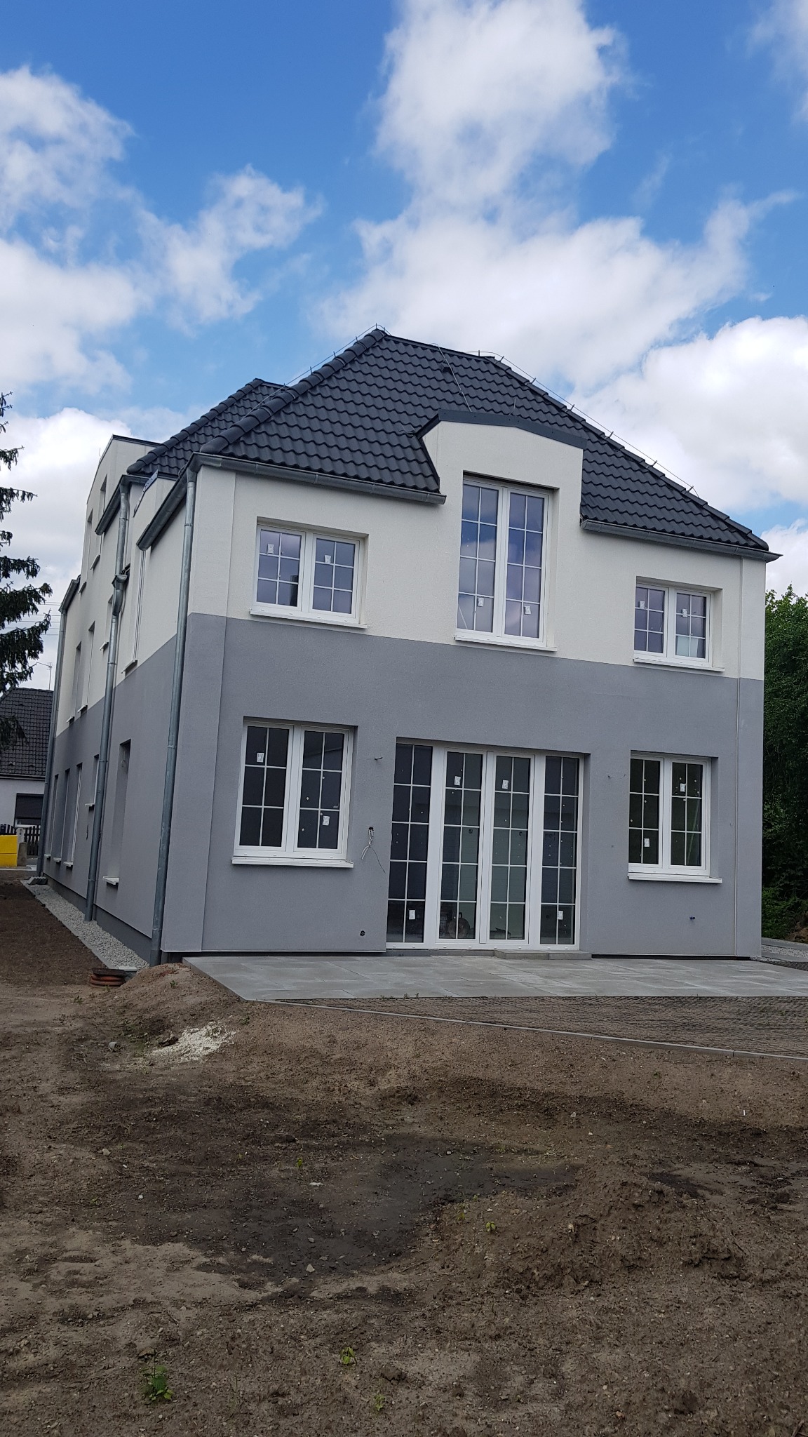 Dwukondygnacyjny dom z szarą dolną częścią i białą górną, z ciemnoszarą dachówką, widoczne duże okna i przeszklone drzwi tarasowe, teren wokół domu w trakcie zagospodarowywania.