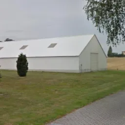 Biały magazyn z blachy falistej z dwuspadowym dachem i świetlikami, otoczony trawnikiem i drzewami iglastymi, widok z chodnika.