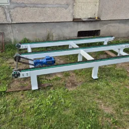 Fragment linii produkcyjnej z napędem łańcuchowym i niebieskim silnikiem elektrycznym, ustawiony na trawie przed budynkiem z szarą elewacją.