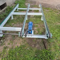 Biała rama przenośnika łańcuchowego z niebieskim silnikiem elektrycznym pośrodku, na tle zielonej trawy i wózka widłowego.