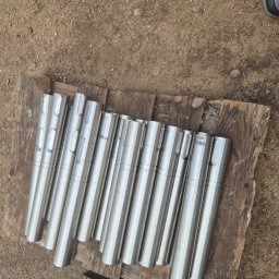 USŁUGI TOKARSKO-ŚLUSARSKIE ARIEL SKUCZYŃSKI - Szereg cylindrycznych, metalowych wałków z rowkiem, ułożonych na drewnianej desce na ziemi.