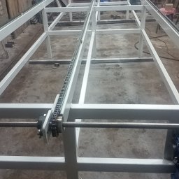 USŁUGI TOKARSKO-ŚLUSARSKIE ARIEL SKUCZYŃSKI - Rama konstrukcyjna z łańcuchem transportowym, wykonana w Malachinie, widoczna w warsztacie z drewnianymi elementami w tle.