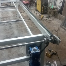 USŁUGI TOKARSKO-ŚLUSARSKIE ARIEL SKUCZYŃSKI - Fragment konstrukcji metalowej z napędem łańcuchowym i silnikiem elektrycznym, widoczne narzędzia i spawarka w tle.