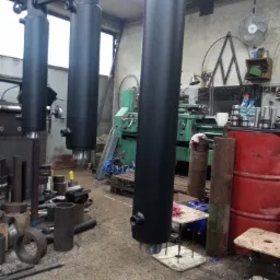 Trzy pomalowane na czarno cylindry hydrauliczne zawieszone na metalowej konstrukcji w warsztacie, w tle widoczna zielona tokarka i metalowe elementy.