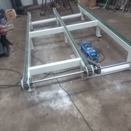 USŁUGI TOKARSKO-ŚLUSARSKIE ARIEL SKUCZYŃSKI - Biała, metalowa rama z łańcuchami transportowymi i niebieskim silnikiem elektrycznym na betonowej podłodze warsztatu.