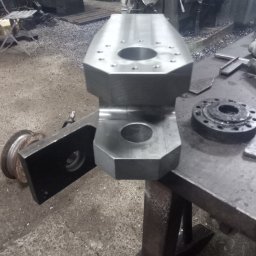 USŁUGI TOKARSKO-ŚLUSARSKIE ARIEL SKUCZYŃSKI - Surowy, metalowy element z otworami, obrobiony na tokarce, leżący na warsztatowym stole; w tle widoczne maszyny i metalowe wióry.