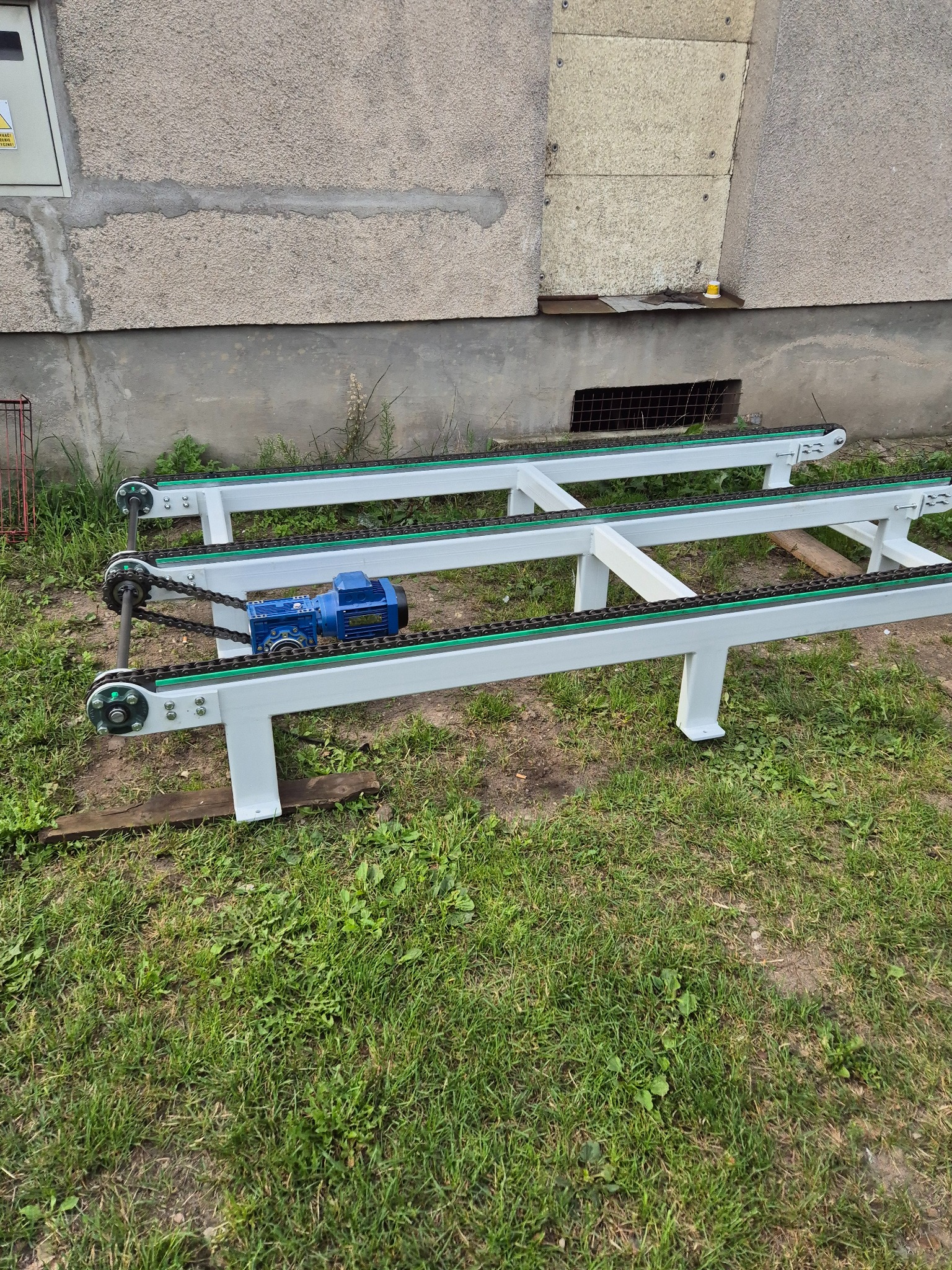 Fragment linii produkcyjnej z napędem łańcuchowym i niebieskim silnikiem elektrycznym, ustawiony na trawie przed budynkiem z szarą elewacją.