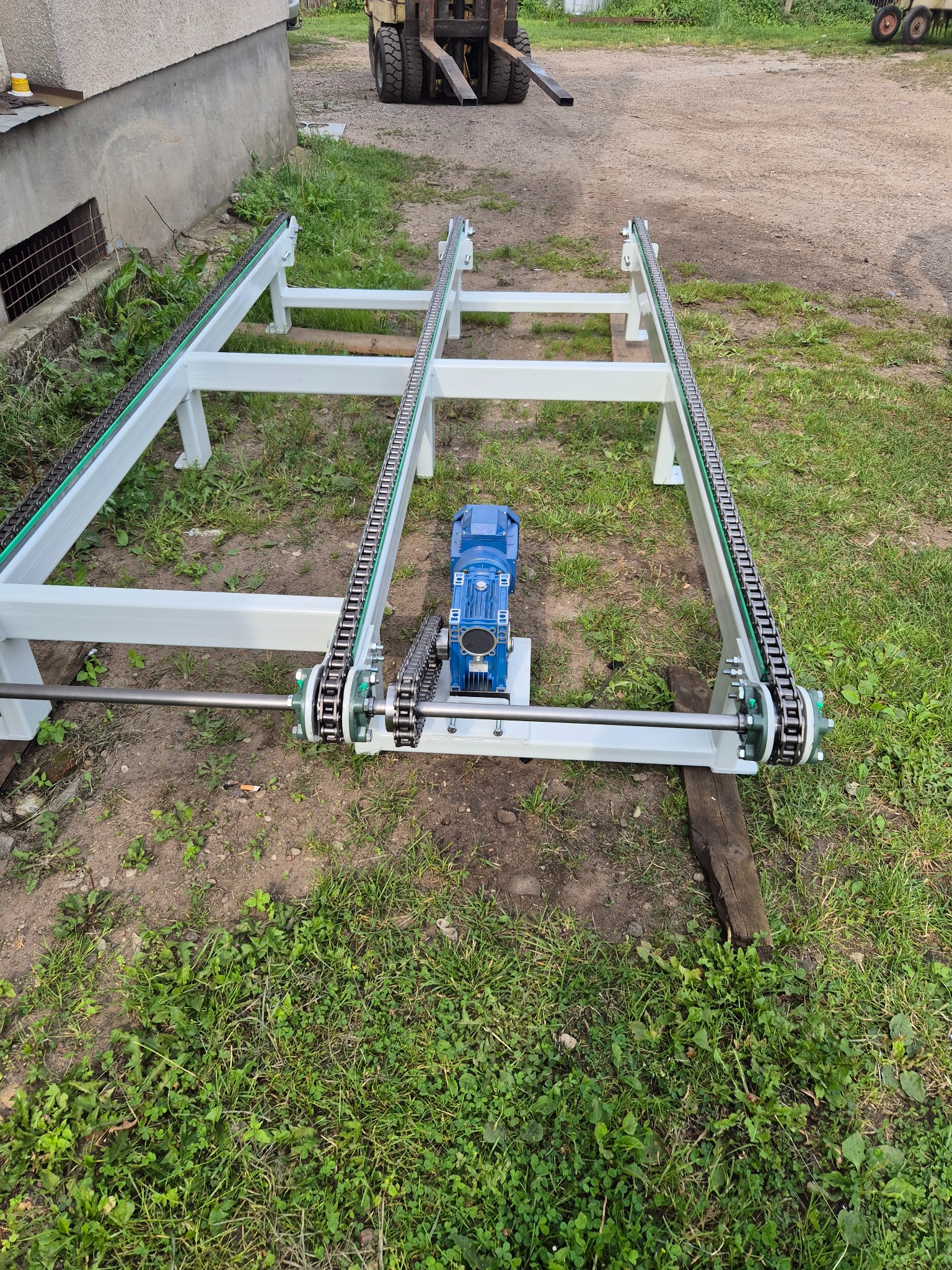 Biała rama przenośnika łańcuchowego z niebieskim silnikiem elektrycznym pośrodku, na tle zielonej trawy i wózka widłowego.