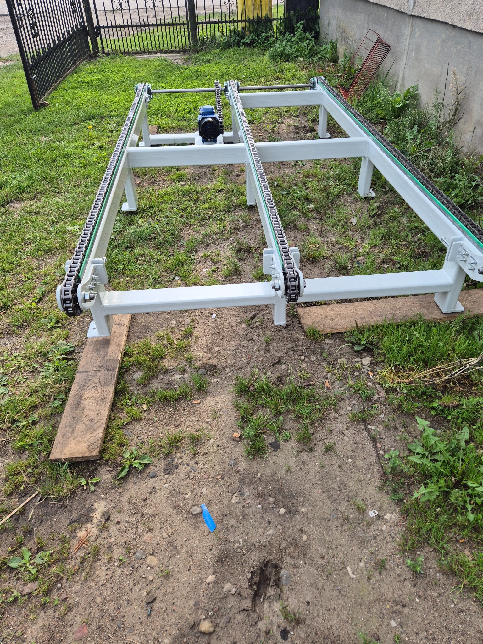 Biała metalowa rama konstrukcji z napędem łańcuchowym i silnikiem elektrycznym, ustawiona na deskach na ziemi z trawą i chwastami, widok z góry.
