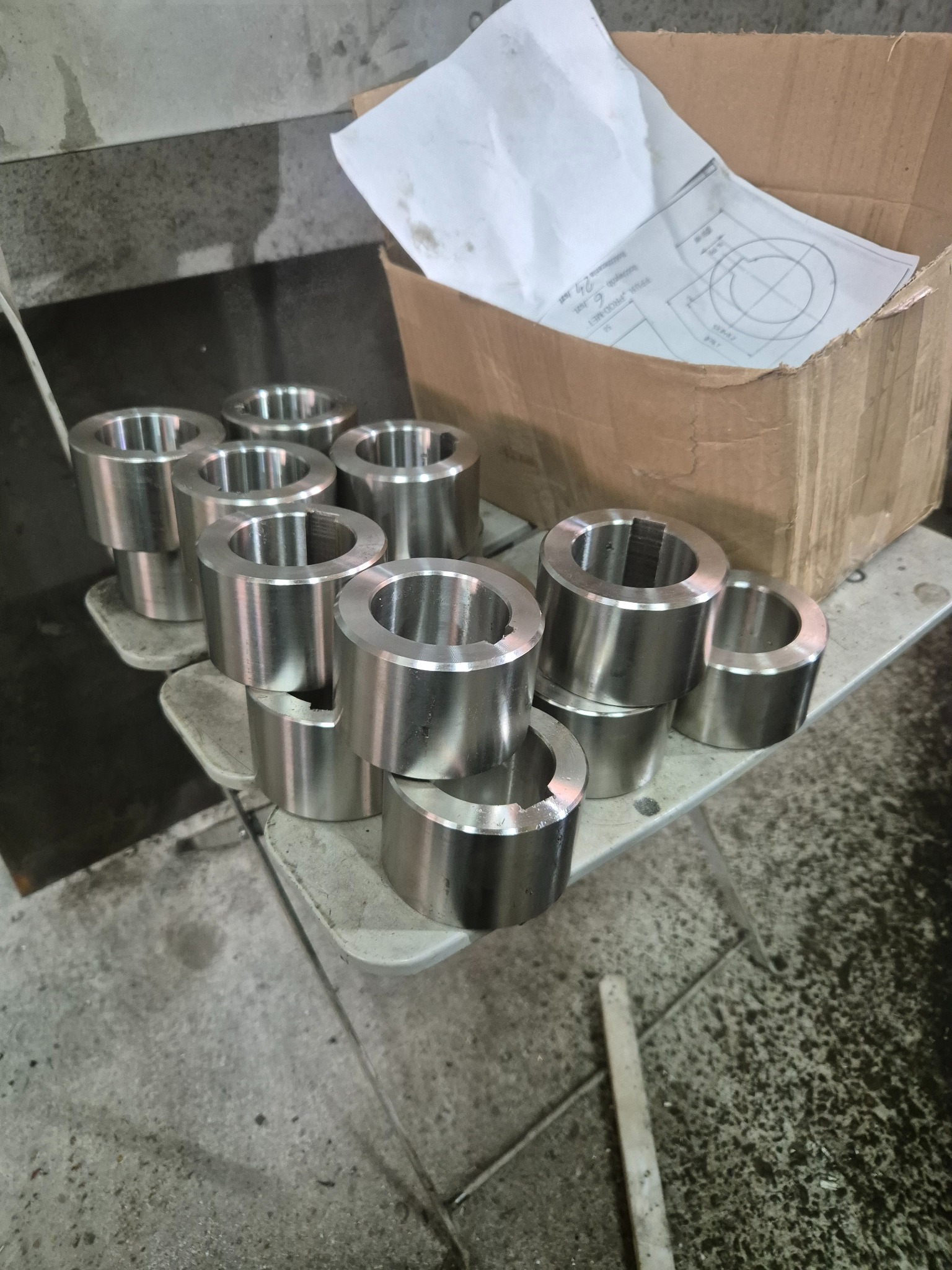 Partia obrobionych, metalowych tulei z widocznym rysunkiem technicznym w kartonowym pudełku w tle, leżących na warsztatowym stole.