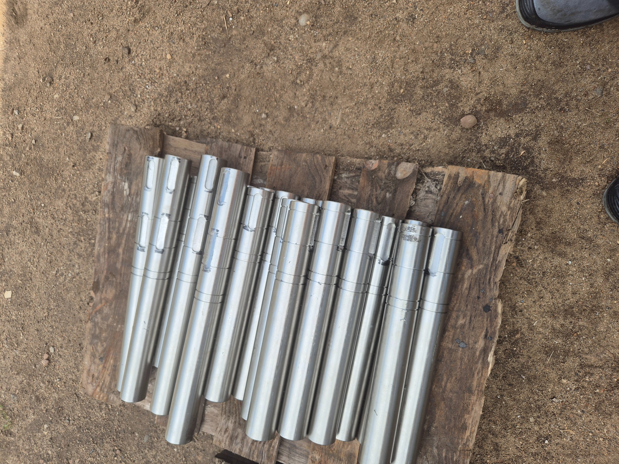 Szereg cylindrycznych, metalowych wałków z rowkiem, ułożonych na drewnianej desce na ziemi.
