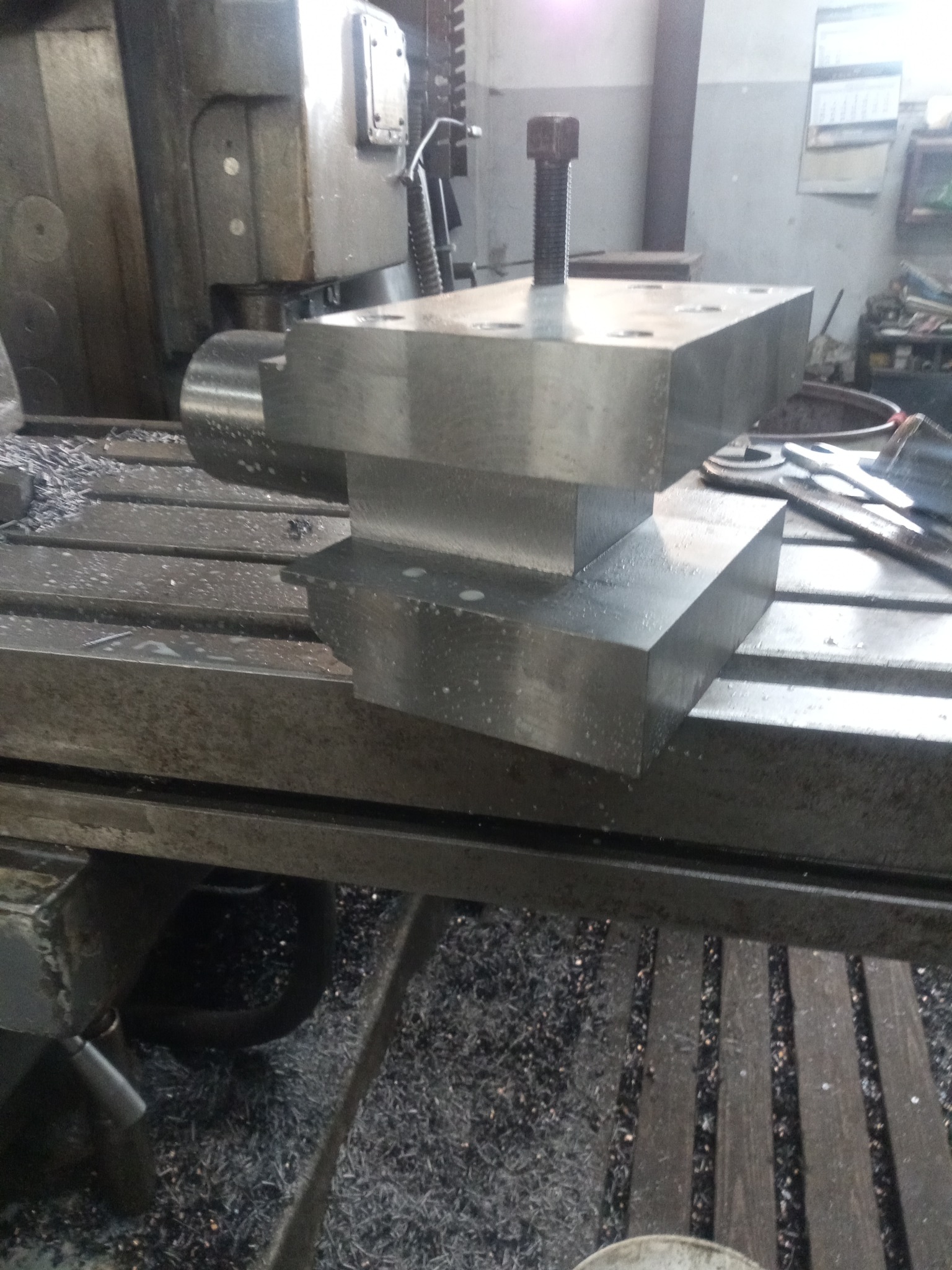 Surowy blok metalu, obrobiony na frezarce, z widocznymi otworami i śrubą, leżący na stole maszyny w otoczeniu wiórów.