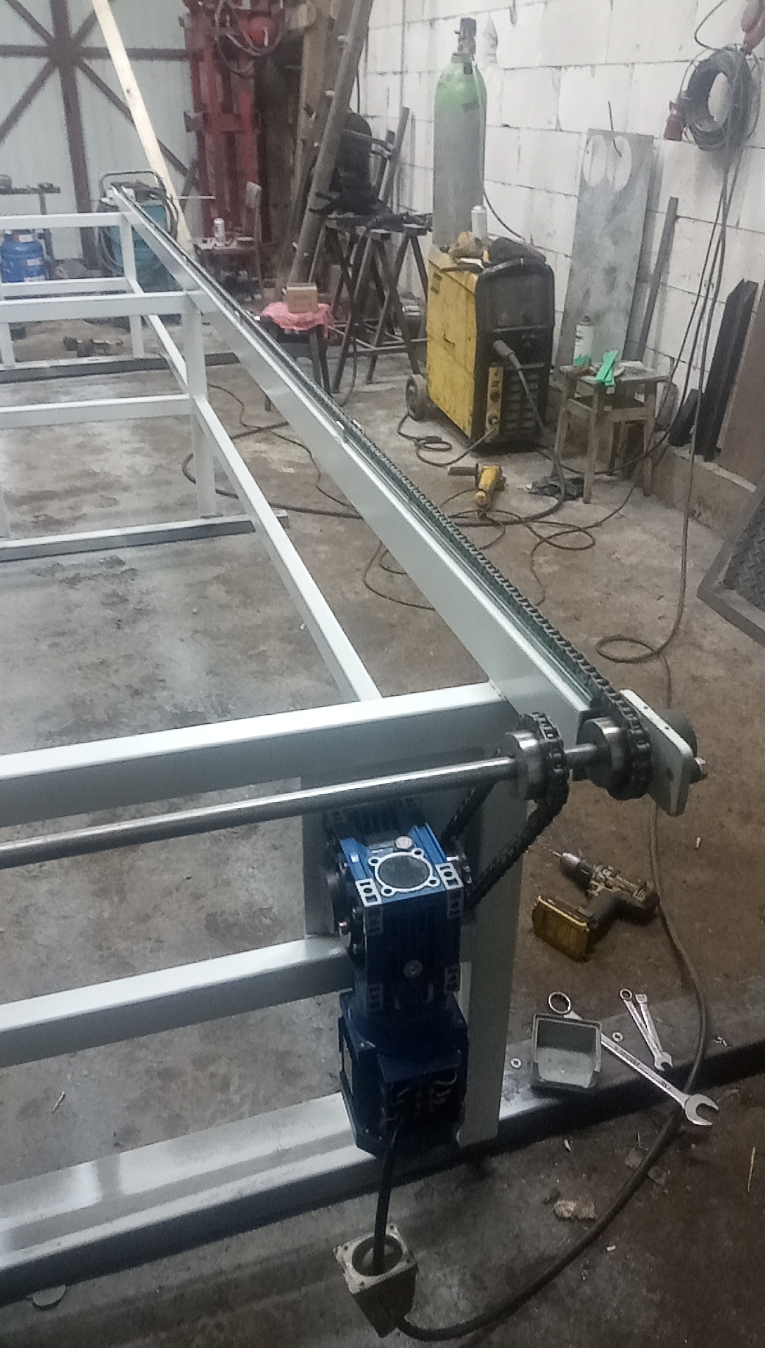 Fragment konstrukcji metalowej z napędem łańcuchowym i silnikiem elektrycznym, widoczne narzędzia i spawarka w tle.