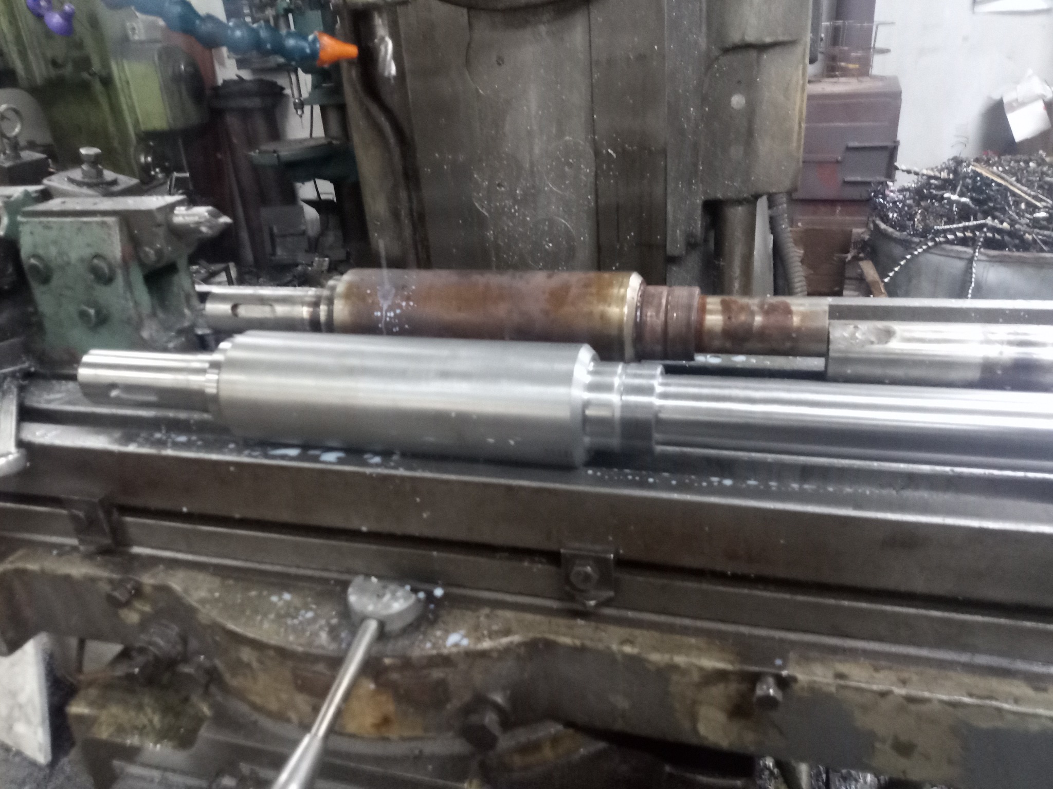 Dwa metalowe wałki o różnej obróbce leżące na stole tokarki, jeden surowy, drugi obrobiony, w tle wióry i maszyny.