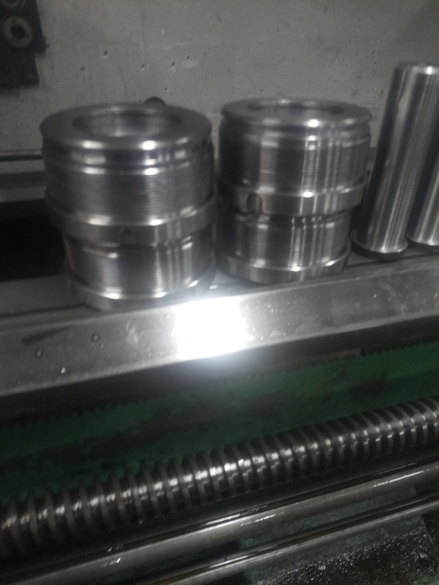 Dwa metalowe elementy cylindryczne z obróbki skrawaniem, leżące na metalowej powierzchni tokarki z widoczną śrubą pociągową.