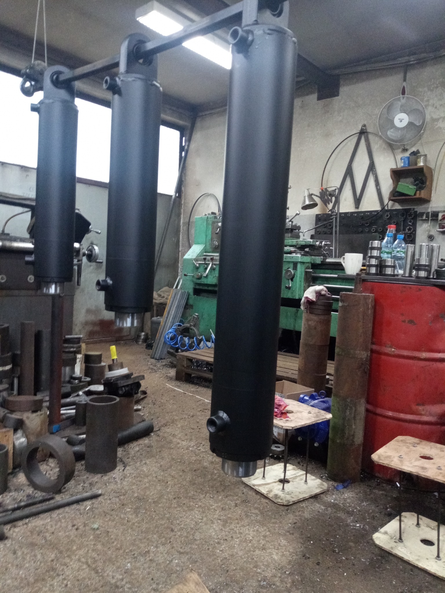 Trzy pomalowane na czarno cylindry hydrauliczne zawieszone na metalowej konstrukcji w warsztacie, w tle widoczna zielona tokarka i metalowe elementy.