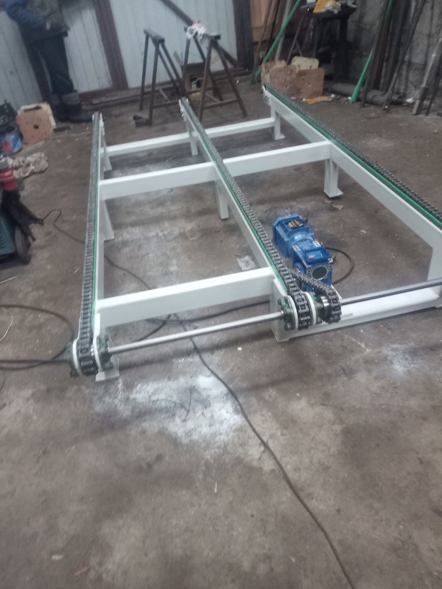 Biała, metalowa rama z łańcuchami transportowymi i niebieskim silnikiem elektrycznym na betonowej podłodze warsztatu.