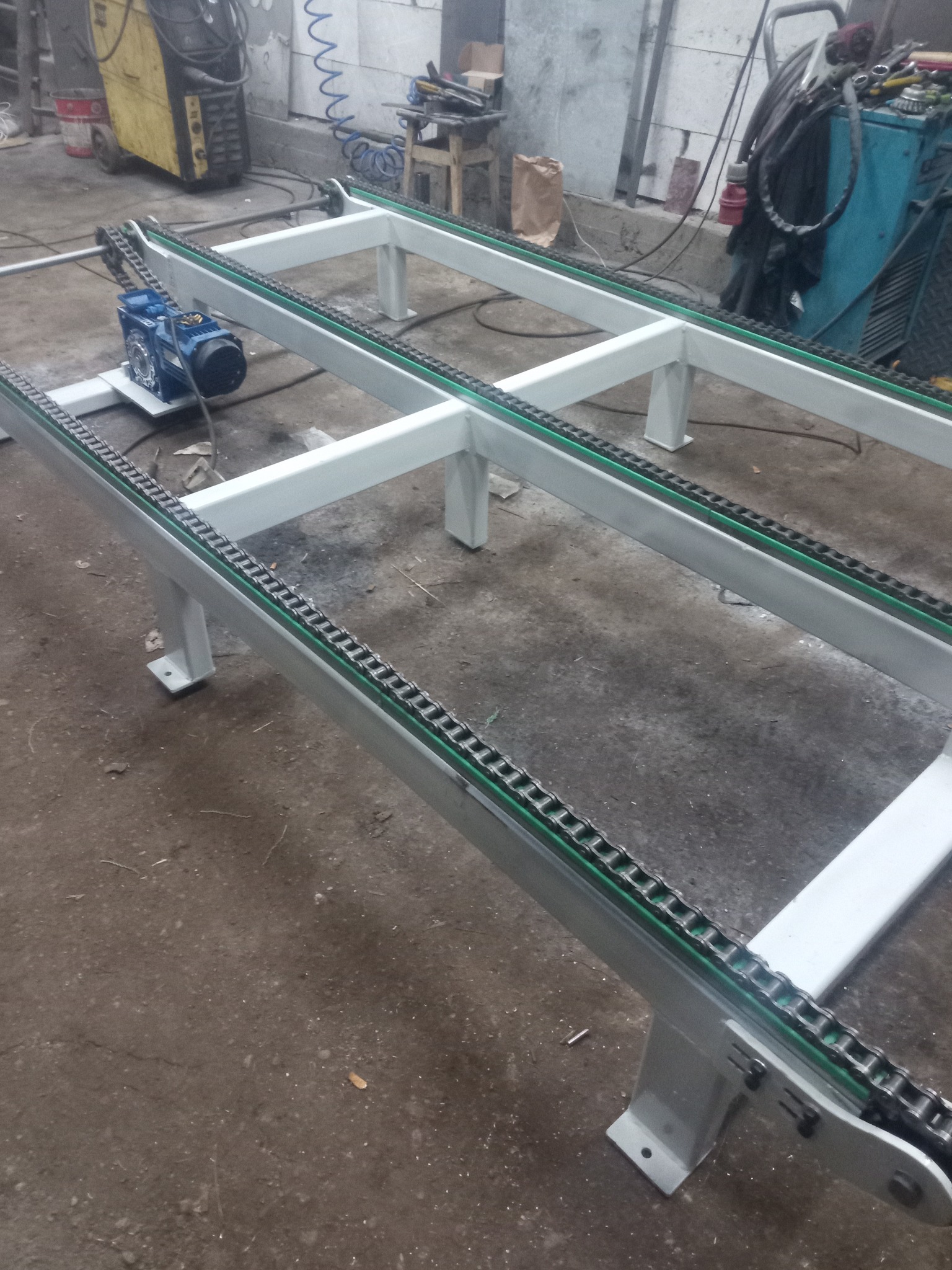 Metalowa konstrukcja przenośnika łańcuchowego z silnikiem elektrycznym, widoczne spawane elementy ramy i łańcuch transportowy