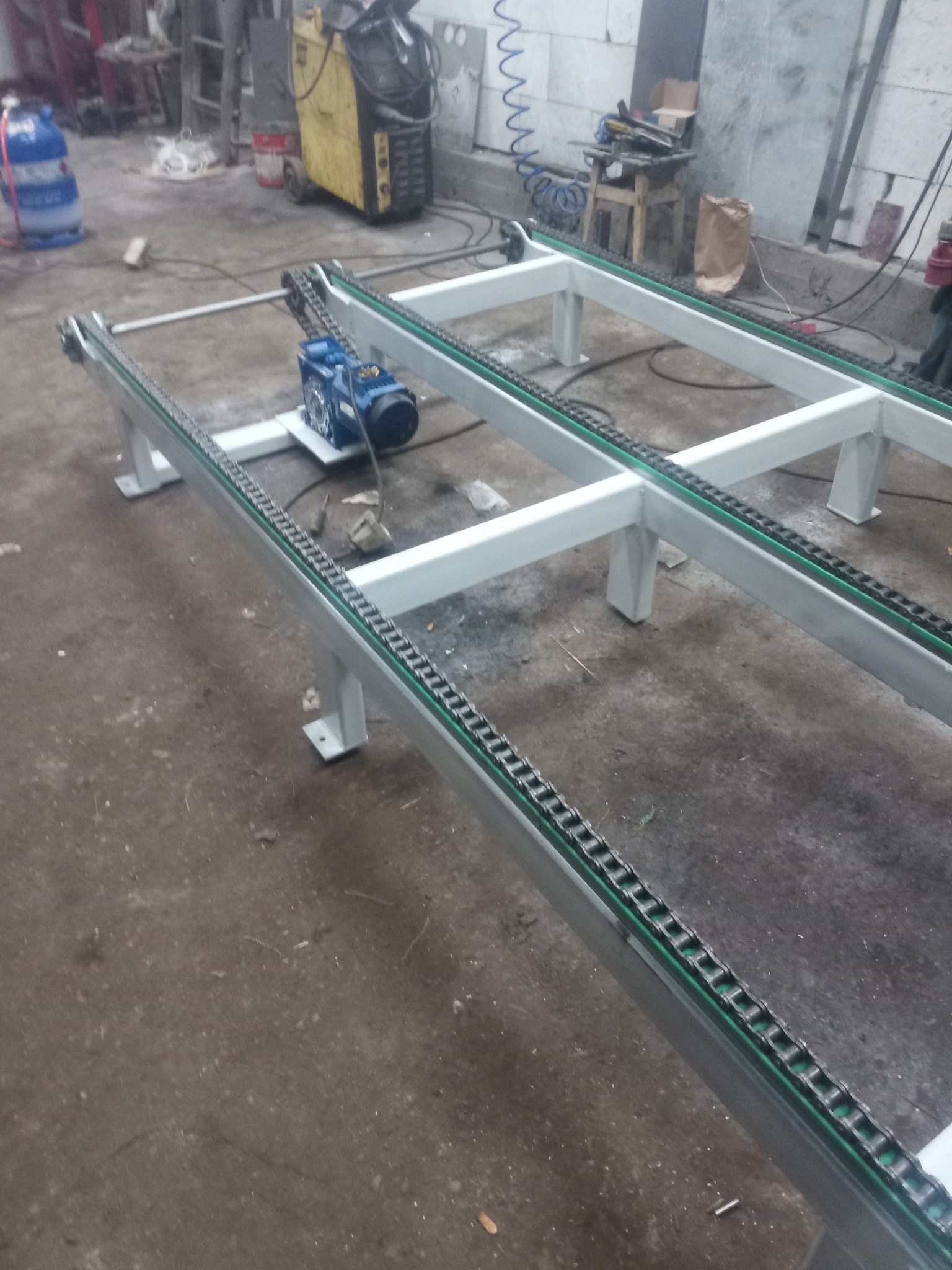 Rama przenośnika łańcuchowego z napędem elektrycznym, na szarym betonowym podłożu warsztatu, widoczne spawane łączenia konstrukcji.