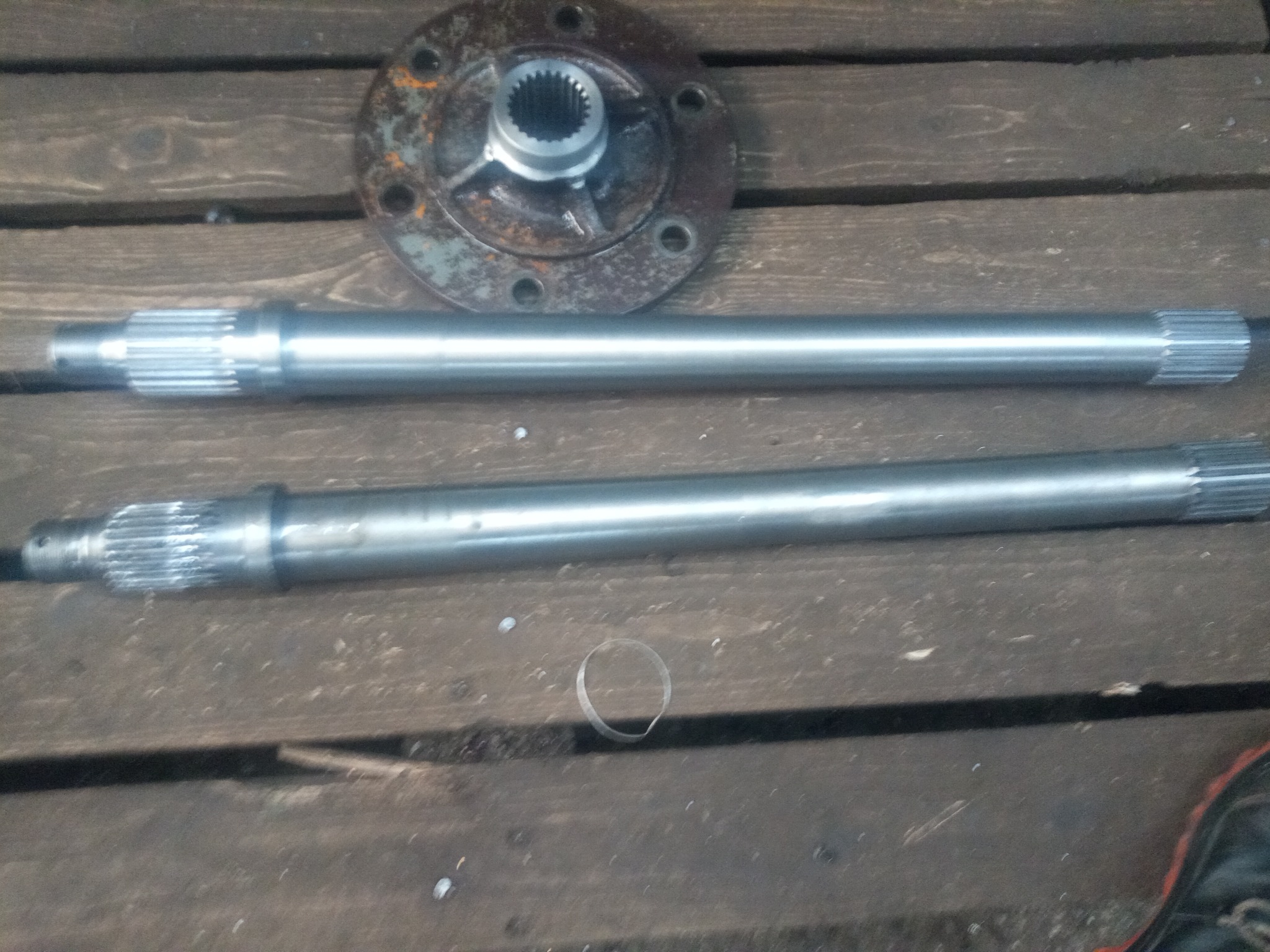 Dwa stalowe wały z zębatymi końcówkami i metalowa flansza z otworami na śruby leżące na drewnianym podłożu z widocznym pierścieniem i fragmentem buta w rogu.