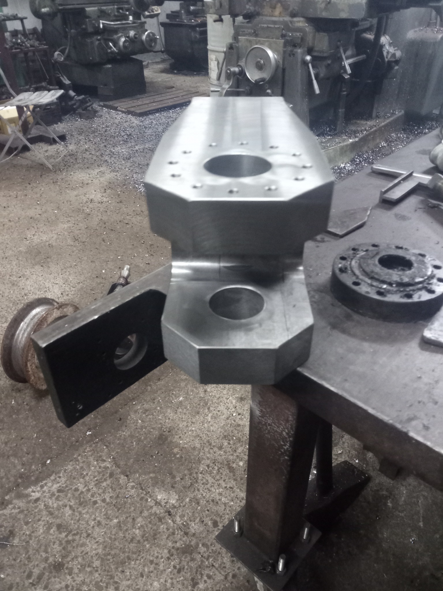 Surowy, metalowy element z otworami, obrobiony na tokarce, leżący na warsztatowym stole; w tle widoczne maszyny i metalowe wióry.