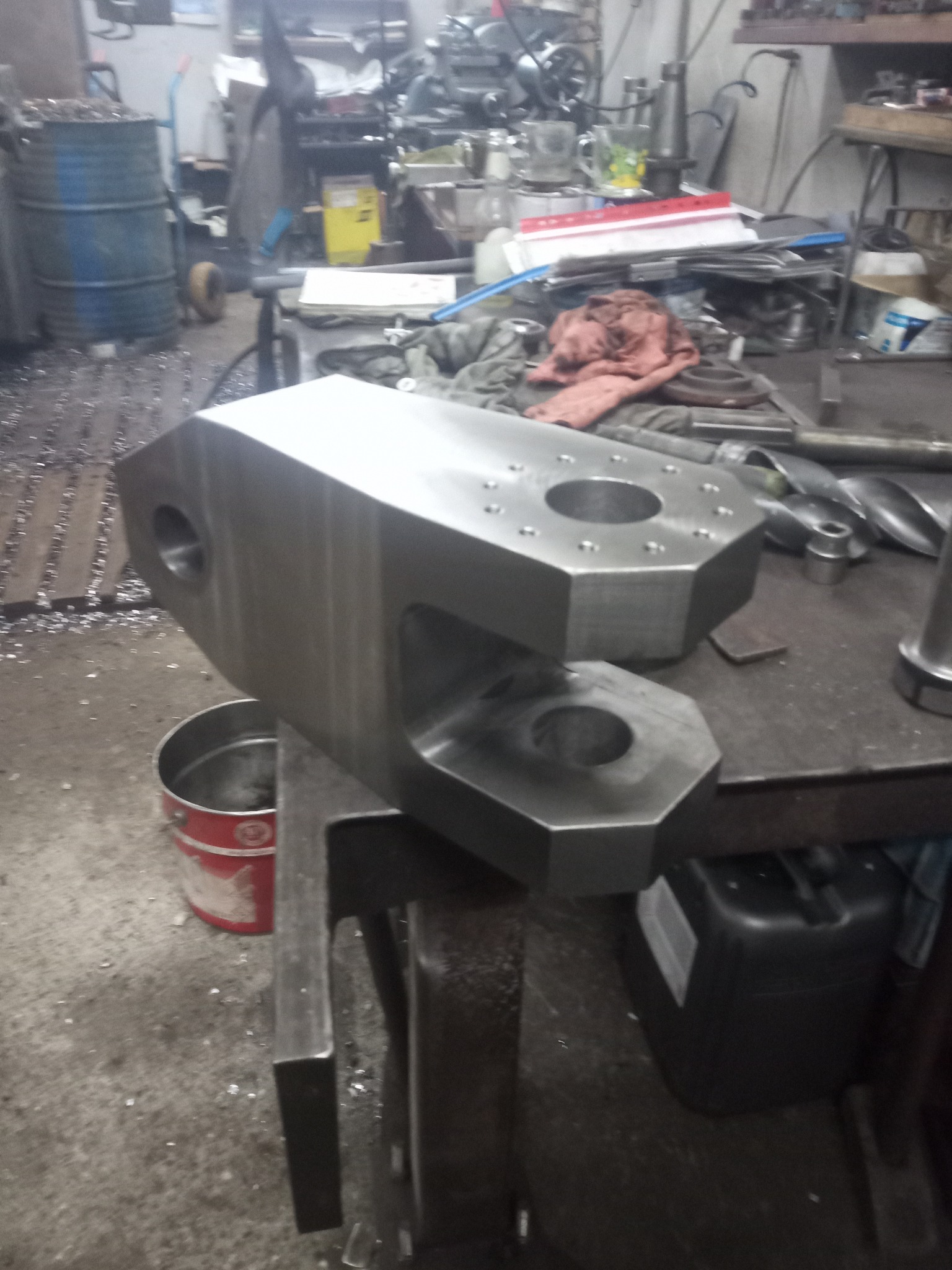 Surowy, obrobiony element metalowy z otworami na warsztatowym stole, w tle widoczne maszyny i narzędzia, wióry na podłodze.