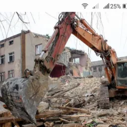 Pomarańczowa koparka na gruzowisku rozbieranego budynku mieszkalnego, widoczne fragmenty ścian i okien, pochmurne niebo w tle.