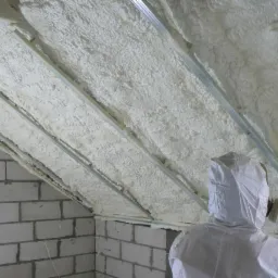 Pracownik w kombinezonie ochronnym aplikuje białą pianę izolacyjną na skośny dach poddasza, widoczne metalowe profile i surowe ściany z białego betonu komórkowego.
