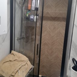 Interfam Tomasz Abramowicz - Kabina prysznicowa z czarną ramą i wzorzystymi płytkami w jodełkę na ścianie. Na półce w kabinie widoczne kosmetyki. Obok kosz z ręcznikami.